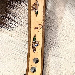 May include: A light-colored leather keychain with a dark brown border. The keychain features three small, hand-painted duck illustrations with green and blue accents. It has a black metal clasp and two silver rivets.