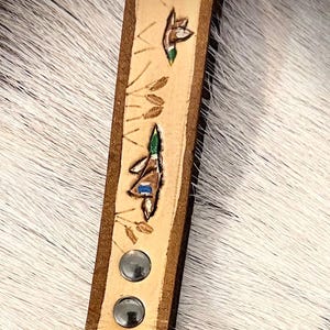 May include: A tan leather keychain strap with a brown border, featuring hand-painted ducks and wheat stalks. The strap is secured with two silver rivets and a black metal clasp.