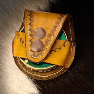 May include: A handcrafted leather coin purse in shades of tan and brown, featuring decorative scalloped edges and two brown snap closures. The purse holds a green and gold coin, showcasing a blend of rustic charm and functionality.