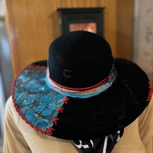 May include: A wide-brimmed black felt hat with a turquoise and black patterned brim. The hat has a red and white trim and a red stitched edge.