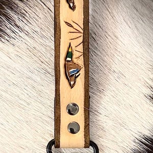 May include: A light-colored leather keychain with a carved duck design. The keychain features two ducks with blue, brown, and green details. The leather is secured with two silver rivets and a black clasp.