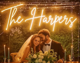 May include: A glowing yellow neon sign displaying the text The Harpers in a cursive script font, mounted above a wooden table with a floral centerpiece, two lit candles, and a wine glass, set against a backdrop of dark green foliage and string lights.