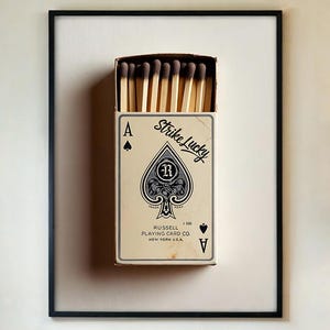 May include: A framed artwork depicting a matchbox designed as the Ace of Spades playing card. The matchbox is cream with black text and a spade design. The text reads "Strike Lucky" and "Russell Playing Card Co, New York, USA".