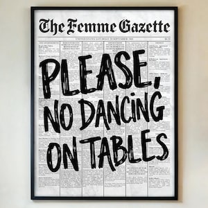 Puede incluir: Un estampado enmarcado con un fondo de estilo periódico. El texto negro en negrita dice "PLEASE, NO DANCING ON TABLES." El titular dice "The Femme Gazette." El estampado está en un marco negro y cuelga de una pared beige.