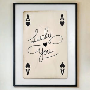 May include: Framed wall art featuring a playing card design. The card is an Ace of Spades with the words "Lucky You" in elegant script. The black frame complements the neutral tones of the artwork, creating a stylish home decor piece.