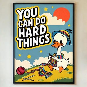 May include: A framed art print featuring a cartoon goose knitting a colorful scarf. The artwork has a blue sky background with a red sun, clouds, and stars. The text "YOU CAN DO HARD THINGS" is displayed in bold, yellow letters.