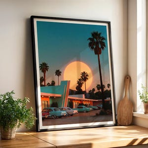 May include: Framed art print featuring a retro motel scene with vintage cars parked in front. The artwork showcases a sunset with a large sun, palm trees, and a teal and orange color palette. The print is in a black frame.