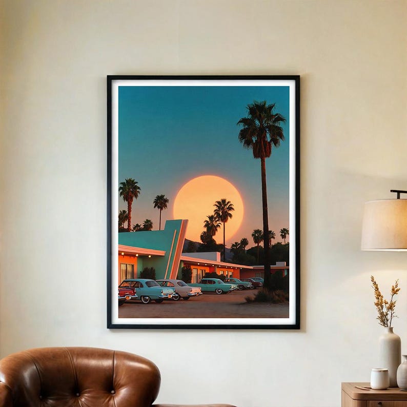 May include: Framed art print depicting a retro motel scene at sunset. The artwork features a large orange sun, palm trees, and vintage cars parked in front of a teal and orange building. The sky is a gradient of blue and orange.