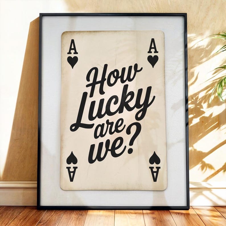 Op de afbeelding: Ingelijste kunstdruk met een ontwerp van een schoppen aas speelkaart. De kaart heeft de tekst "How Lucky are we?" in zwart schrift. De print staat tegen een cr&egrave;mekleurige achtergrond en is ingelijst in een zwart frame.