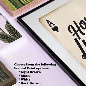 Op de afbeelding: Ingelijste kunstdruk met een ontwerp van een harten aas speelkaart met de tekst "Home is where the heart is". De print wordt gepresenteerd in een zwart frame, met opties voor lichtbruine, zwarte, witte en donkerbruine frames.