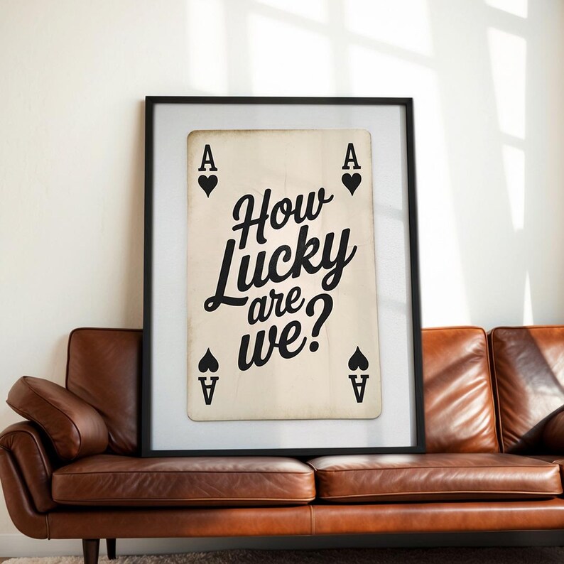 Op de afbeelding: Een ingelijste print met een speelkaartontwerp. De kaart is een Aas van Schoppen met de tekst "How Lucky are we?" in zwart schrift. De print zit in een zwarte lijst en wordt weergegeven boven een bruine leren bank.