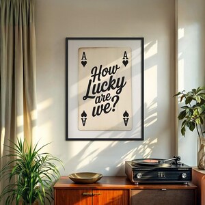 Op de afbeelding: Een ingelijste print met een speelkaart met de Aas van Schoppen symbolen en de tekst "How Lucky are we?" in een stijlvol lettertype. De print wordt weergegeven boven een houten kast met een platenspeler en een decoratieve kom.