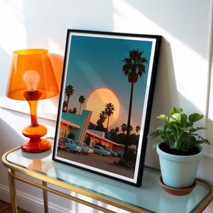 May include: A framed art print depicting a retro scene with palm trees, vintage cars, and a large sun. The print is in a black frame and is displayed on a glass-topped table next to an orange lamp and a potted plant.