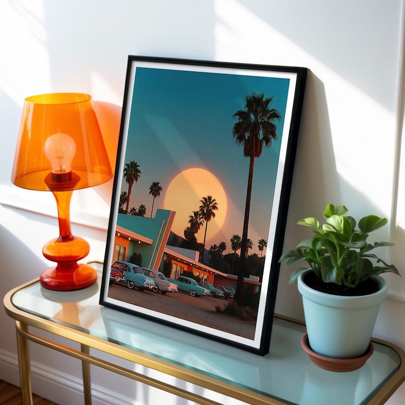 May include: A framed art print depicting a retro scene with palm trees, vintage cars, and a large sun. The print is in a black frame and is displayed on a glass-topped table next to an orange lamp and a potted plant.