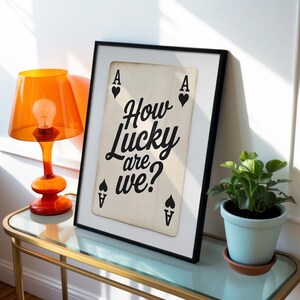 Op de afbeelding: Een ingelijste print met een speelkaartontwerp en de tekst "How Lucky are we?" in zwart schrift. De Aas van Schoppen is zichtbaar. Een oranje lamp en een potplant staan ook in de afbeelding.