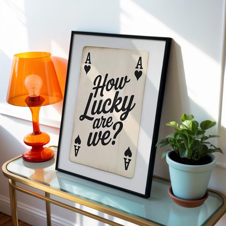 Op de afbeelding: Een ingelijste print met een speelkaartontwerp en de tekst "How Lucky are we?" in zwart schrift. De Aas van Schoppen is zichtbaar. Een oranje lamp en een potplant staan ook in de afbeelding.