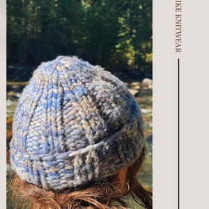 May include: A blue and gray knit beanie with a classic fisherman style. The beanie is worn by a person with red hair, standing in front of a forest and a river.