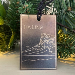 May include: A brown leather ornament with a gold outline of a cliff and the text "HA LING" on the front. The ornament is hanging from a black cord.