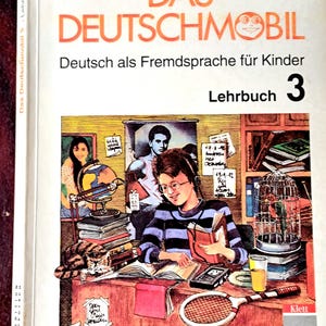 Pode incluir: Livro didático "DAS DEUTSCHMOBIL" para alemão como língua estrangeira para crianças. A capa apresenta uma ilustração de um menino lendo, um globo e uma raquete de tênis. Publicado por Klett e Mass Media.