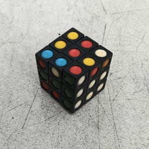 Könnte beinhalten: Ein schwarzer 3x3 Würfel-Puzzle mit farbigen Kreisen auf jeder Seite. Die Farben sind rot, gelb, blau und weiß.