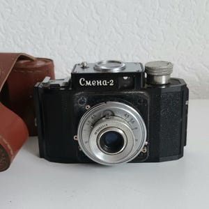 Pode incluir: Uma câmera de filme de 35 mm preta e prateada com o texto "Смена-2" na parte superior. A câmera tem uma lente com um anel prateado e uma capa de couro preta.