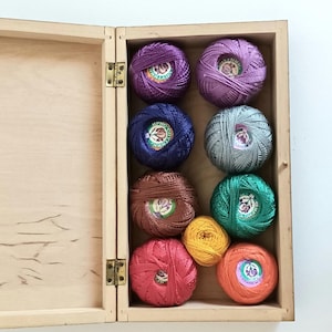 Può includere: Una scatola di legno con un coperchio incernierato, aperta per rivelare una collezione di rocchetti di filo da ricamo colorati. I rocchetti sono disposti a griglia e includono sfumature di viola, blu, verde, arancione, rosso e marrone.