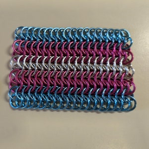 Puede incluir: Una pequeña pieza rectangular de cota de malla en colores azul, rosa y plateado. La cota de malla está hecha de pequeños anillos de metal entrelazados.