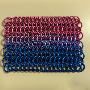 May include: A small, rectangular piece of chainmail in the colors of the bisexual pride flag. The chainmail is made of small, interlocking metal rings. The top band is pink, the middle band is purple, and the bottom band is blue.