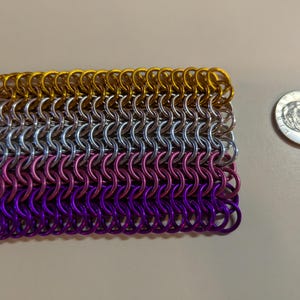 May include: A rectangular piece of chainmail composed of gold, silver, pink, and purple rings. A US quarter is next to the chainmail for size comparison. The chainmail is made of small, interlocking metal rings.