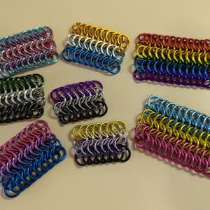 Op de afbeelding: Een verzameling van zeven kleine, rechthoekige kettingpostvlaggen in verschillende kleuren, waaronder roze, blauw, geel, groen, zwart, wit en zilver. De vlaggen zijn gemaakt van met elkaar verbonden metalen ringen.