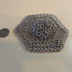 May include: A hexagonal piece of chainmail made of small, interlocking metal rings. A silver quarter coin is next to the chainmail for size comparison. The chainmail is silver and has a textured surface.