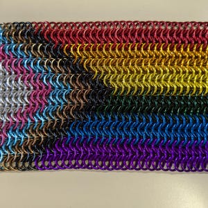 May include: A rectangular piece of chainmail composed of small, colorful metal rings. The colors include red, orange, yellow, green, blue, purple, pink, silver, and black, arranged in horizontal stripes. The chainmail is likely a decorative or functional item.