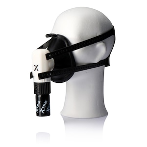 Puede incluir: Una mascarilla facial en blanco y negro con un sistema de correas, exhibida en una cabeza de maniquí blanca. La mascarilla tiene un componente blanco con un logotipo "X" negro y una pieza cilíndrica negra con texto "XTRM".