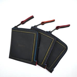 Puede incluir: Tres carteras cuadradas negras con ribetes y cremalleras de colores. Cada cartera tiene un ribete de color diferente: rojo, amarillo y azul. Las cremalleras tienen un tirador negro con un acento de color. Las iniciales "MHI" están grabadas en la parte delantera de una cartera.