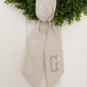 May include: A beige and white striped fabric bow with a green embroidered letter "G" hanging on a green wreath.