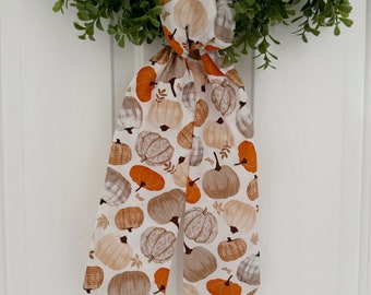 May include: A white fabric hair scarf with a pattern of orange and brown pumpkins. The scarf is tied in a bow and hangs from a wreath.