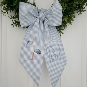 May include: A blue and white gingham bow with an embroidered stork carrying a baby and the text "It's a Boy!"