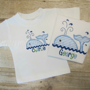 Op de afbeelding: Twee witte T-shirts met blauwe en witte gestreepte walvis-borduurwerk. De walvis op het linker T-shirt heeft de naam "Charlie" eronder geborduurd. De walvis op het rechter T-shirt heeft de naam "George" eronder geborduurd.