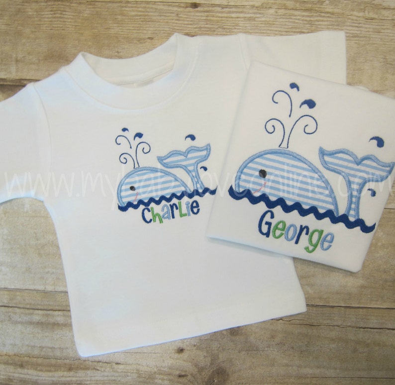 Op de afbeelding: Twee witte T-shirts met blauwe en witte gestreepte walvis-borduurwerk. De walvis op het linker T-shirt heeft de naam "Charlie" eronder geborduurd. De walvis op het rechter T-shirt heeft de naam "George" eronder geborduurd.