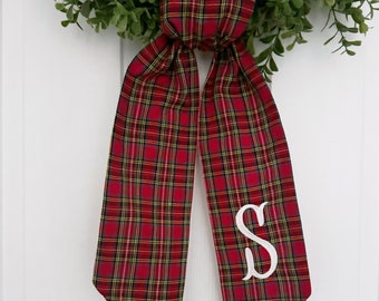 May include: A red and green plaid bow with a white letter "S" monogrammed on it. The bow is tied around a green wreath.