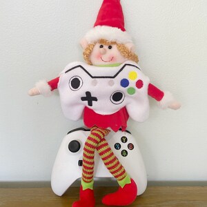 May include: A plush elf doll wearing a white and red outfit with a video game controller design. The elf is sitting on a white video game controller.