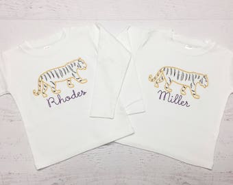 May include: Two white long-sleeved shirts with embroidered tiger designs. The tigers are outlined in gold with black stripes. One shirt has the name "Rhodes" and the other "Miller" in purple script.