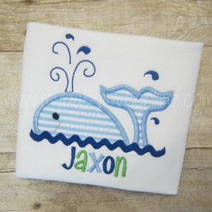 Op de afbeelding: Een witte romper met een blauwe walvis geborduurd erop. De walvis is blauw-wit gestreept en heeft een blauwe staart. De walvis blaast water uit zijn spuitgat. De romper heeft de naam "Jaxon" geborduurd in groene letters.