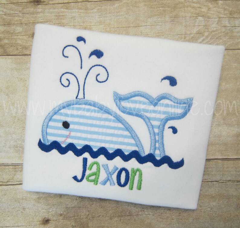 Op de afbeelding: Een witte romper met een blauwe walvis geborduurd erop. De walvis is blauw-wit gestreept en heeft een blauwe staart. De walvis blaast water uit zijn spuitgat. De romper heeft de naam "Jaxon" geborduurd in groene letters.