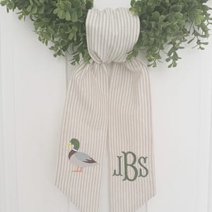 May include: A green wreath with a striped ribbon featuring a duck illustration and the initials "JBS" in green. The ribbon is beige with vertical gray stripes, adding a touch of rustic charm to the wreath.