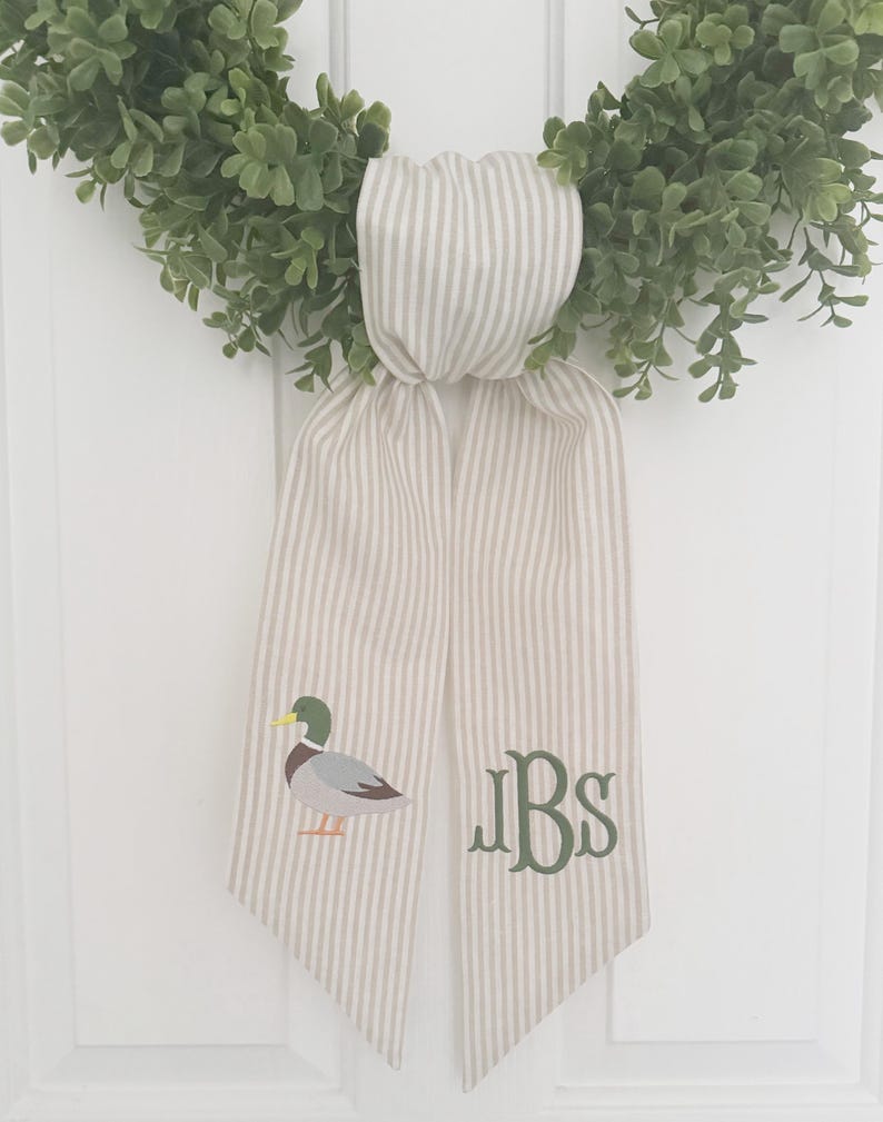 May include: A green wreath with a striped ribbon featuring a duck illustration and the initials "JBS" in green. The ribbon is beige with vertical gray stripes, adding a touch of rustic charm to the wreath.