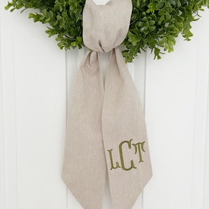 May include: A beige linen wreath bow with a green embroidered monogram "LCT" on the front.