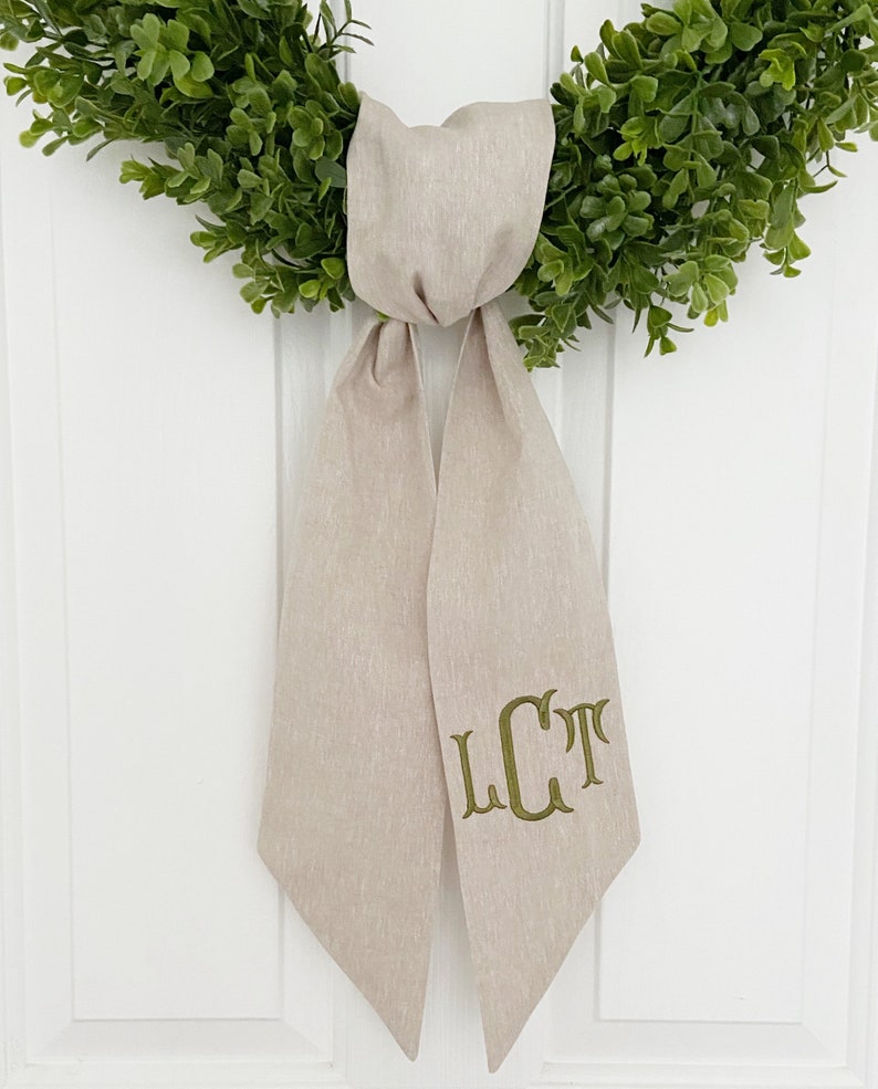 May include: A beige linen wreath bow with a green embroidered monogram "LCT" on the front.