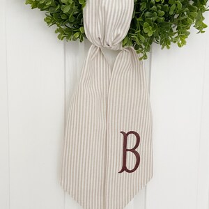May include: A beige and white striped fabric bow with a burgundy embroidered letter "B" hanging on a white door.