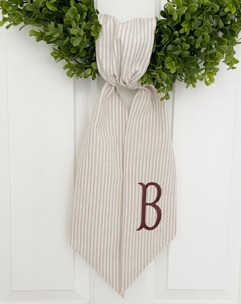 May include: A beige and white striped fabric bow with a burgundy embroidered letter "B" hanging on a white door.